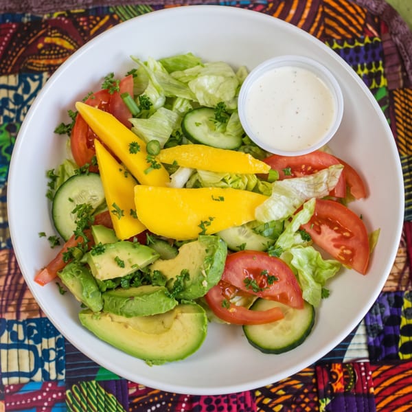 Fresh salad with lettuce, tomatoes, cucumbers, avocado, mango slices, and a side of dressing.