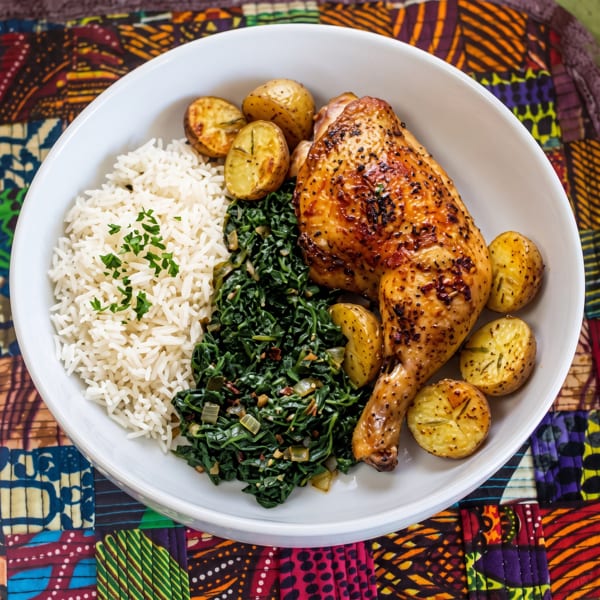 A white bowl filled with roasted chicken, rice, sautéed greens, and baby potatoes on a colorful placemat.
