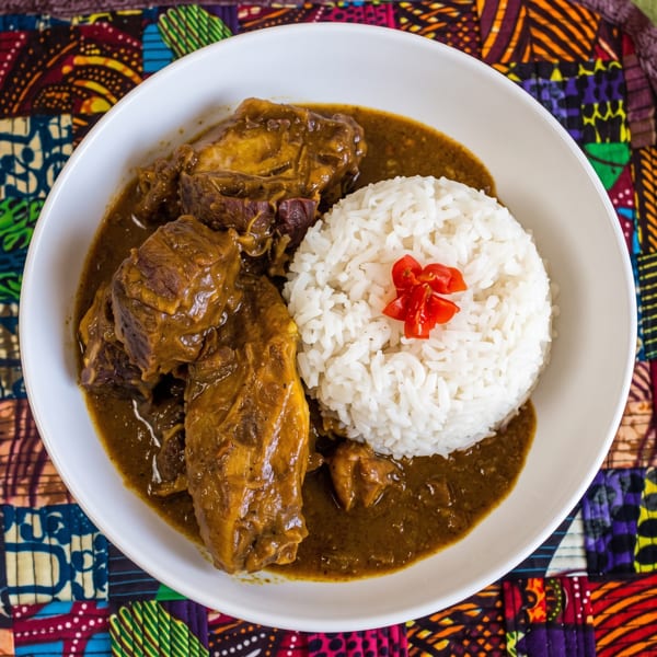 A white bowl of West African stew with meat and rice, garnished with red pepper.