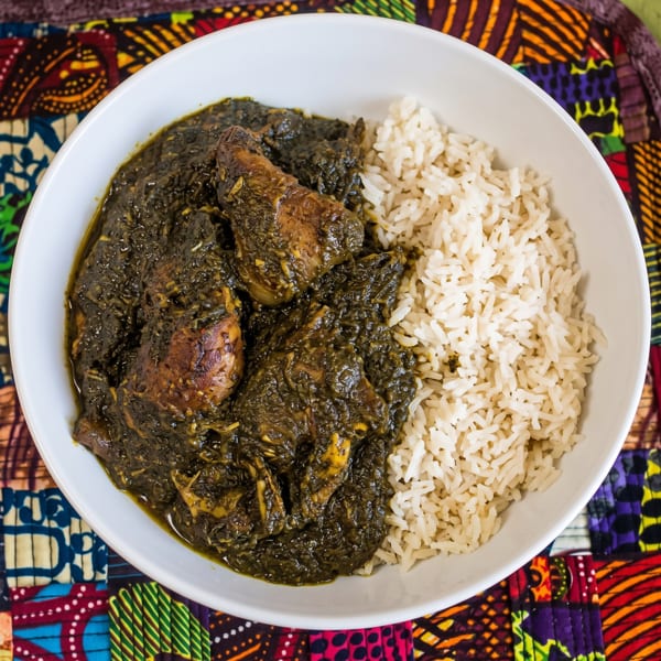 A white bowl filled with dark green stew and chicken, alongside white rice, on a colorful patterned mat.